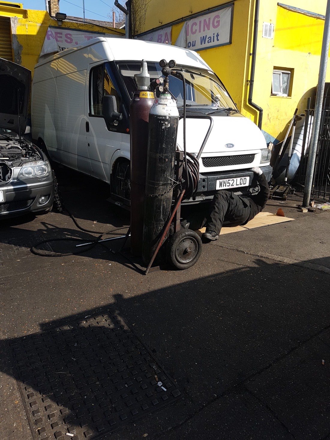 A mechanic working underneath a white Ford Transit van in the Ranic Motors yard, with tools and gas bottles alongside.