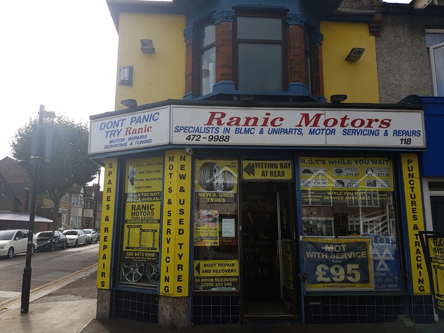 The yellow and blue Ranic Motors shopfront at 118 High Street South, East Ham, with hand-painted signs for MOTs, new and used tyres, servicing and repairs.