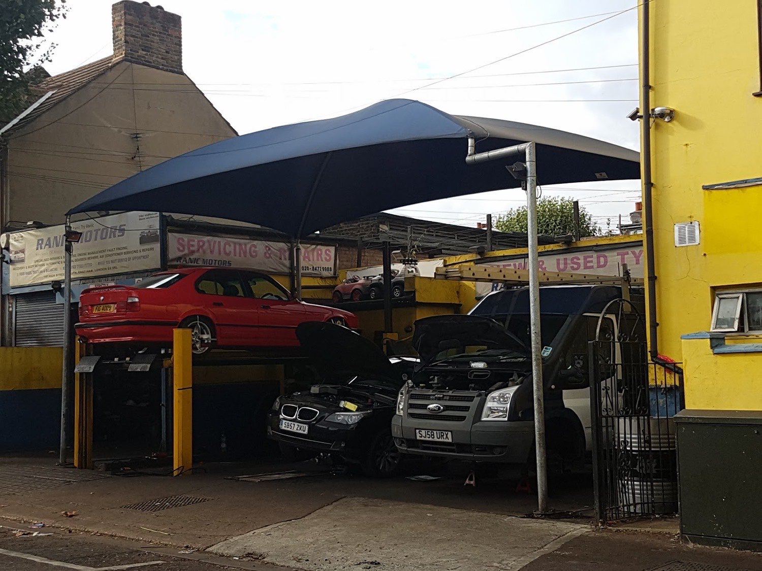 Cars raised on a ramp and others with bonnets up under the covered work bay at the rear of Ranic Motors in East Ham.